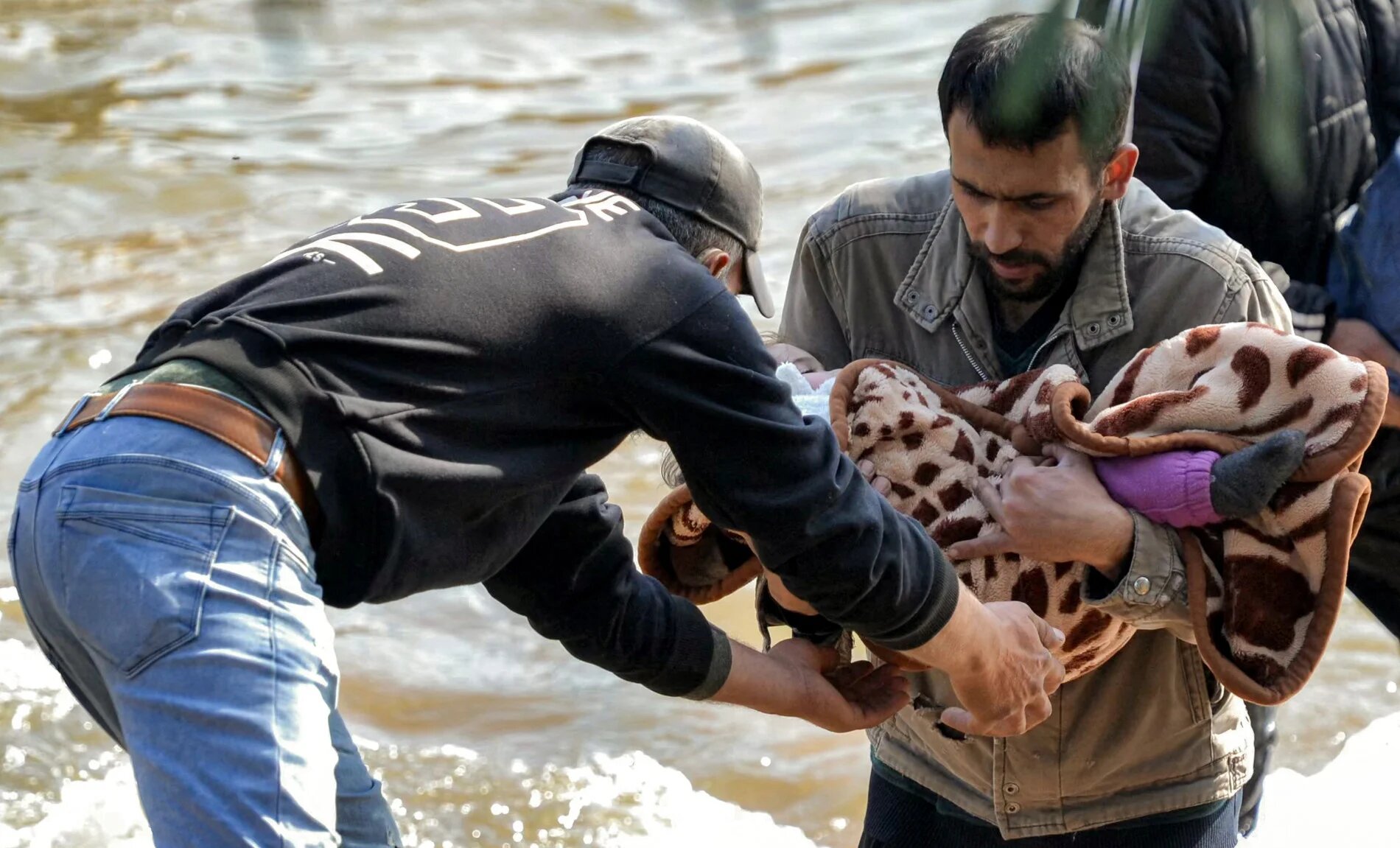 A man receives a blanketed child from another crossing the Nahr al-Kabir river, forming the border between Syria's western coastal province and northern Lebanon in the Hekr al-Daher area on 11 March 2025 (AFP)