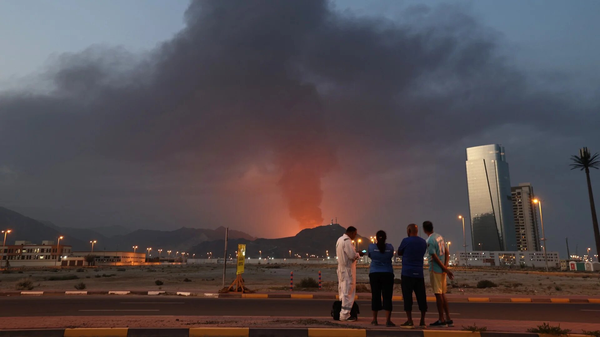 Foreign workers look at a tall plume of black smoke rising after an explosion in the Fujairah industrial zone in the UAE on 3 March 2026 (Fadel Senna/AFP)